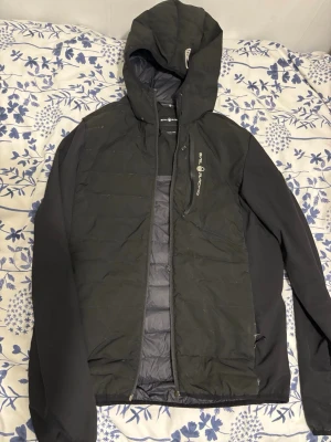 Svart pufferjacka med huva från Sail Racing. - Svart pufferjacka från Sail Racing med huva och dragkedja framtill. Jackan har quiltade partier på framsidan och en bröstficka med dragkedja. Perfekt för kyliga dagar och har en sportig look.      OBS!!! Det är fel med dragkedjan på jackan så det går inte att stänga jackan.