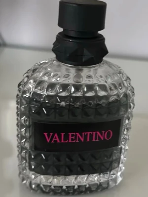 Valentino parfymflaska  - Snygg parfym från Valentino i en genomskinlig glasflaska med nitar och svart etikett med rosa text. Flaskan har en unik, kantig design och svart kork. Perfekt för dig som gillar lyxiga och trendiga dofter.