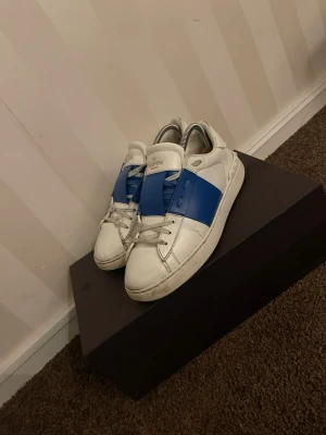 Valentino vita sneakers med blå detalj - Säljer ett par vita sneakers från Valentino med en bred blå rem över vristen. Skorna har klassisk låg profil, rund tå och vita snören. Tillverkade i läder med gummisula och diskret logga på plösen. Perfekta för dig som gillar stilrena men ändå unika sneakers.