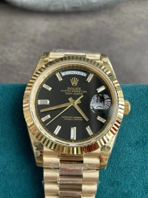 Rolx Day-Date guld klocka svart urtavla - Exklusiv Rolx Oyster Perpetual Day-Date med svart urtavla och dag- samt datumvisning. Klockan har en elegant guldfärgad boett och länkarmband, samt markerade index i silver. Klassisk räfflad bezel och tydlig logotyp vid tolvslaget.                                                        En lasergraverad logotyp är placerad vid klockan sex, och på boettens baksida finns en stämplad logotyp i varje hörn. Mekanisk klocka. ✅Brand new! Fast shipping 