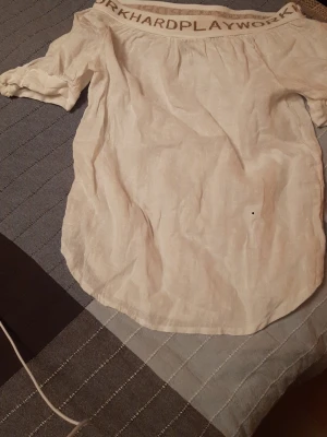 Offshoulder blus med textdetalj - Säljer en vit offshoulder blus med beige textdetalj runt halsen där det står 'WORKHARDPLAY'. Blusen har korta ärmar och är gjord i ett lätt, luftigt tyg som känns som linne. Perfekt för en avslappnad och trendig look. Utan storlek label, så mått på bilderna. Jag tror är det storlek L/XL.