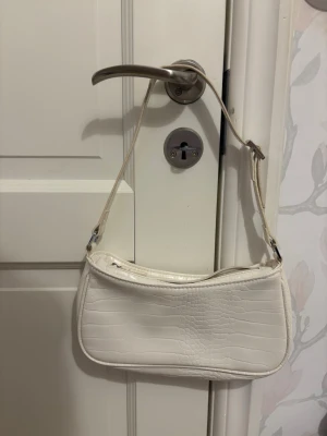 Vit axelväska  - Stilren vit handväska med justerbar axelrem och krokodilpräglat mönster. Väskan har en avlång, mjukt rektangulär form och stängs med dragkedja. Perfekt för dig som gillar trendiga accessoarer med lite extra edge. Väskan har ett stort fack. Skriv gärna innan ni köper❤️