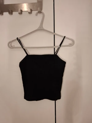 Svart ribbad linnetopp med smala band - En svart ribbad linnetopp med smala axelband och croppad passform. Materialet känns mjukt och stretchigt, vilket gör den bekväm att bära.