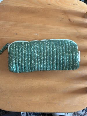 Trendiga Systrarna grene Grön quiltad pennskrin med blommönster - Säljer en grön quiltad pennskrin med små vita blommor och dragkedja. Plånboken är av tyg och har en avlång, mjuk form som gör den perfekt för kort, mynt eller småsaker. Enkel och stilren design som sticker ut med sitt mönster.från systrarna grene