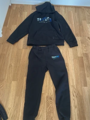 Svarta mjukisbyxor från Thrasher - Säljer ett par svarta mjukisbyxor från Thrasher med tryckt logga i vitt och blått på ena benet. Byxorna har resår i midjan och muddar vid bensluten, perfekta för chill dagar eller streetwear-stil.