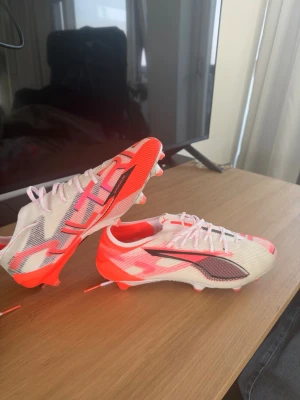 Puma vita och röda fotbollsskor - Säljer ett par snygga fotbollsskor från Puma i vitt med röda och svarta detaljer. Skorna har låg profil, snörning och mönstrad ovandel i syntetmaterial för bra grepp och kontroll på planen. Perfekta för dig som vill sticka ut på fotbollsplanen.