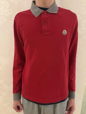 Röd långärmad piké från Moncler - Snygg röd långärmad pikétröja från Moncler med grå krage.. Tillverkad i mjuk bomull som känns skön mot huden. Perfekt för dig som gillar klassisk men ändå sportig stil.