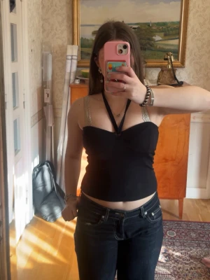 Svart croppad halterneck topp - Snygg svart croppad halterneck topp med twistad detalj framtill. Toppen är ribbad och har en omlottkänsla över bysten. Perfekt till jeans eller kjol för en trendig look.