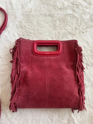 Röd mockaväska med fransar från Maje - Cool röd handväska i mocka från Maje med fransar längs sidorna och rektangulärt handtag i rött läder. Väskan har en fyrkantig form och ger en trendig boho-vibe. Perfekt accessoar för att lyfta din outfit.