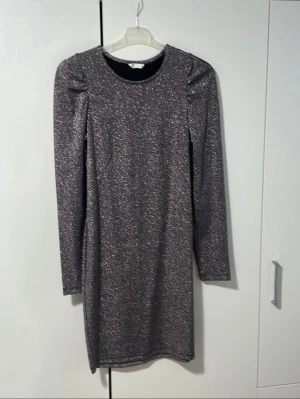 Glittrig långärmad klänning - Snygg glittrig klänning i mörkgrått med lång ärm och rund halsringning. Rak passform och axelvaddar för markerad siluett. Perfekt för dig som vill sticka ut med en stilren look.