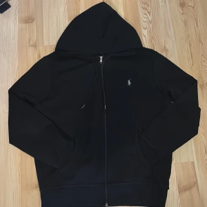 Svart hoodie från Ralph Lauren - Svart hoodie från Ralph Lauren med klassisk logga på bröstet. Tröjan har huva med snörning, dragkedja framtill och två fickor. Tillverkad i mjuk bomullsmix och har långärmad passform. Perfekt för chill dagar och enkel att matcha med jeans eller joggers. Använd 4 gånger sen jag köpte den för ungefär 5 månader sen. Så den är nästan helt oanvänd. Pris kan diskuteras!