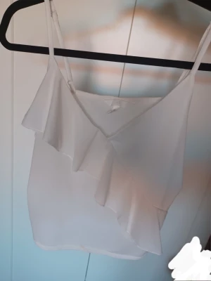 Vit volang topp🫰🏻🫰🏻💞 - Säljer en söt vit linnetopp med smala axelband och volangdetalj framtill. Toppen är lite genomskinlig. Den har en liten sminkfläck. Kommer inte till användning🫰🏻🫰🏻