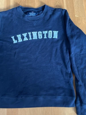 Marinblå sweatshirt från Lexington - Klassisk marinblå sweatshirt från Lexington med tryckt logga i vitt över bröstet. Tröjan har rund halsringning och långa ärmar. Tillverkad i mjukt bomullsmaterial som är skönt att bära. Perfekt för en avslappnad och stilren look.