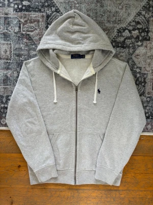 Ljusgrå Polo Ralph Lauren Zip Up Hoodie - Storlek M - Hej! 👋 Säljer en klassisk ljusgrå Polo Ralph Lauren zip-up hoodie i storlek M med marinblå pony-logga. 🐎✨ Tröjan är i mycket fint skick överlag, men det finns en mindre missfärgning/fläck precis vid dragkedjan (se sista bilden). Den är nästintill osynlig så länge man inte letar efter den. Garanterat 100% äkta med originaltvättlappar kvar (ingen QR-kod på denna årsmodell). Ett perfekt premium-basplagg! Fraktar snabbt! 🚛📦💬 