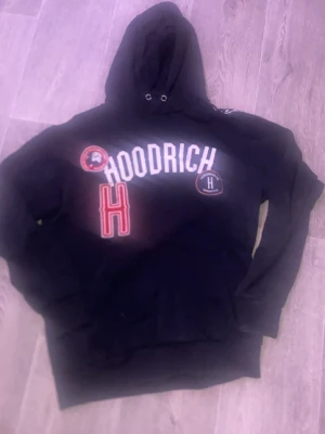 Svart hoodie från Hoodrich - Säljer en svart hoodie från Hoodrich med stor vit logga på bröstet, röd broderad H och två patchar med text och grafik. Klassisk huva med dragsko och känguruficka framtill. Perfekt för dig som gillar streetwear och vill sticka ut.