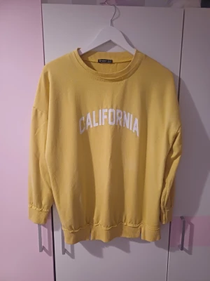 Tröja - Oversized gul sweatshirt med vit text 'CALIFORNIA' på bröstet. Tröjan har rund halsringning, ribbade muddar vid ärmslut och nederkant samt avslappnad passform. Perfekt för en chill och trendig look.