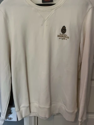 Vit/beige sweatshirt från Morris - Klassisk vit/beige sweatshirt från Morris med broderad logga och text på bröstet. Tröjan har rund halsringning, ribbade muddar och är tillverkad i mjukt material som känns skönt mot huden. Perfekt för en clean och stilren look.