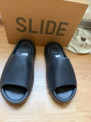 Svarta Adidas Yeezy Slide tofflor - Säljer ett par svarta Adidas Yeezy Slide tofflor, helt oanvända, priset kan diskuteras fråga gärna om fler bilder!