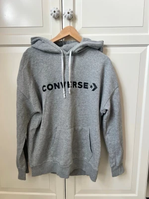 Grå hoodie från Converse - Snygg grå hoodie från Converse med svart logga över bröstet. Klassisk modell med huva och vita dragsnören samt en stor magficka. Tillverkad i mjuk bomullsblandning, perfekt för en avslappnad streetstyle-look.