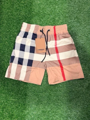 Rutig shorts från Burberry - Säljer ett par klassiska rutiga shorts från Burberry med beige, vit, marinblå och röd färg. Shortsen har elastisk midja med svart snörning och den ikoniska Burberry-rutan. Perfekta för sommaren och riktigt snygga till streetwear.