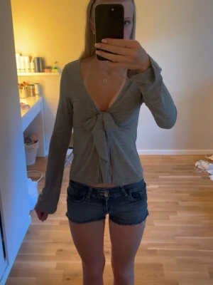 Grå långärmad topp med rosett - Snygg grå långärmad topp med djup v-ringning och stor rosettdetalj framtill. Modellen är croppad och har en mjuk, stretchig känsla. Perfekt för dig som gillar trendiga och feminina detaljer. Kommer inte till användning tvättas innan jag skickar den