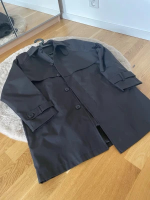 Svart klassisk trenchcoat - Stilren svart trenchcoat med knappar framtill och detaljerade ärmremmar. Jackan har en klassisk krage och vindslå över bröstet. Perfekt för dig som vill ha en tidlös och enkel look. Materialet känns slätt och tåligt.