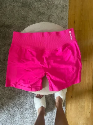 Rosa träningstights shorts från DFYNE - Säljer ett par knallrosa träningstights shorts från DFYNE med hög midja och ribbad linning. Shortsen är tillverkade i ett stretchigt syntetmaterial och har en tight passform som sitter skönt vid träning. Perfekta för gymmet eller löprundan!