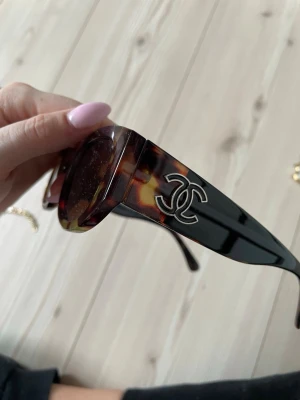 Solglasögon i brun leopard - Snygga solglasögon från Ch med brunmelerad leopardmönstrad båge och bruna glas. Cat-eye formade och med tydlig logga i silver på skalmarna. Perfekt accessoar för att lyfta din stil.