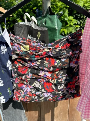 Blommig bandeau linnetopp från Zara - Säljer en linnetopp från Zara med blommigt mönster i rött, lila, grönt och vitt på svart botten. Toppen har tunna axelband och snygga rynkade detaljer framtill. Perfekt för dig som vill ha en färgstark och trendig look.