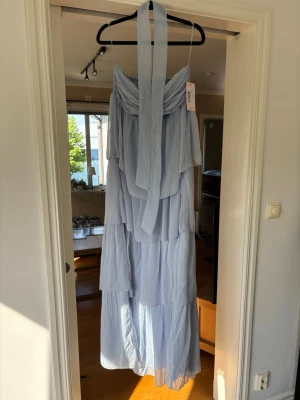 Ljusblå långklänning med volanger - Romantisk ljusblå långklänning med flera lager volanger och smock i ryggen. Klänningen har axelbandslös design med breda band som kan knytas runt halsen. Perfekt för sommarens alla tillfällen.