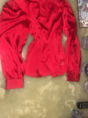 Röd långärmad blus - Trendig röd blus i glansigt tyg med lång ärm. Blusen har en figurnära passform och rund halsringning. Perfekt för dig som vill sticka ut med färg och stil. Dessutom är den helt oanvänd 