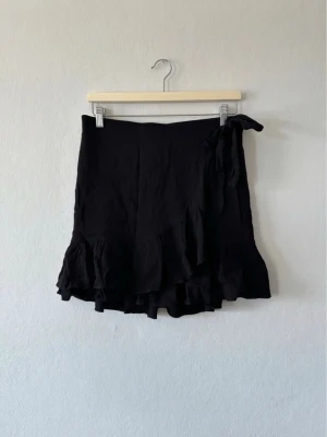 Svart omlottkjol med volang - Snygg svart kort omlottkjol med volangkant och knyt i midjan. Kjolen har en feminin och trendig look, perfekt för att styla med en enkel topp eller stickad tröja. Materialet känns lätt och flowy.