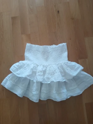 Vit volangkjol med spetsdetaljer strl XS - Supersöt vit kortkjol med bred smock i midjan och dubbla volanger. Kjolen har fina spetsdetaljer och broderier som ger en romantisk känsla. Perfekt för sommarens alla tillfällen och lätt att matcha med olika toppar.
