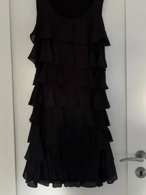 Svart volangklänning med lager - Snygg svart ärmlös klänning med flera lager volanger som ger en cool och lekfull look. Klänningen har rund halsringning och är tillverkad i ett lätt, flödande material som känns bekvämt mot huden.
