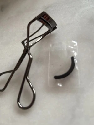 Svart ögonfransböjare med extra dyna - En klassisk ögonfransböjare i metall med svart finish och ergonomiskt handtag. Medföljer en extra svart silikon-dyna i plastförpackning. Perfekt för att skapa snygg böj på fransarna. Passar de flesta ögonformer och är enkel att använda.
