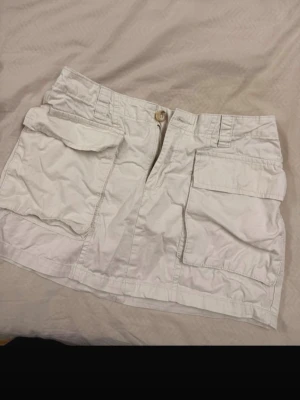 Vit cargoskort med stora fickor - Snygg vit cargoskort med två stora fickor framtill och dragkedja med knapp. Skortsen har bälteshällor och är gjord i ett lätt bomullstyg, perfekt för en avslappnad och trendig look.
