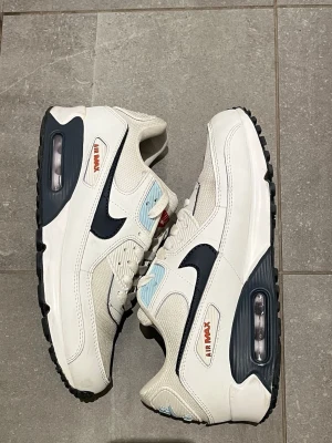 Nike Air Max 90 vita sneakers - Nike Air Max 90 sneakers i vitt med svarta detaljer och ljusblå accenter. Klassisk chunky sula med synlig Air-enhet och meshpaneler för extra andningsförmåga. Röd Nike-logga på plösen och sidan. Perfekta för dig som gillar retrostil och streetwear. Skorna är i strl 43. (Skorna är ej original)