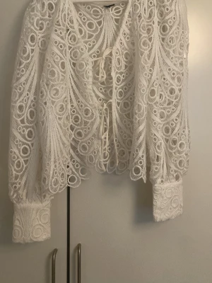 Vit spetsblus/kofta med knytning - Elegant vit kofta/blus i spetsliknande tyg med knytning framtill. Blusen har långa, vida ärmar med breda muddar. Perfekt att styla över ett linne eller topp. Grövre, tjockare material