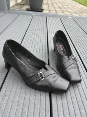 Svarta skor i äkta skinn från Marie strl 39 - Stilrena svarta skor från Marie i äkta skinn med fyrkantig tå och låg klack. Dekorativt metallspänne framtill ger en klassisk look. Perfekta för dig som gillar tidlös design och vill ha ett par bekväma och eleganta skor.