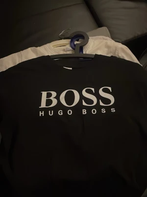 Svart BOSS t-shirt från Hugo Boss - Snygg svart t-shirt från Hugo Boss med stor vit BOSS-logga framtill. Klassisk rund halsringning och korta ärmar. Perfekt för en stilren och trendig look. HELT NY UTAN PRISLAPP