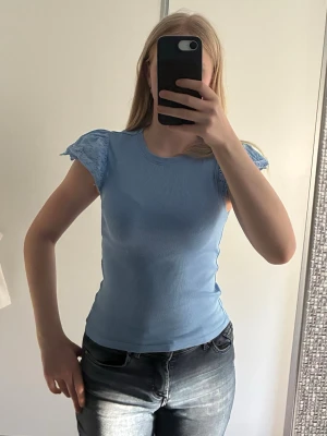 Blå ribbad topp med spetsärmar - Snygg ljusblå topp med ribbad struktur och korta spetsärmar med broderade blommor. Modellen är figurnära och toppen har rund halsringning. Perfekt för dig som vill ha en söt och trendig look.