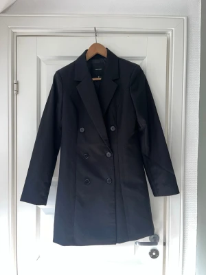 Svart dubbelknäppt kappa Vero Moda  - Stilren svart kappa från Vero Moda med dubbelknäppning och klassisk krage. Lång modell med raka ärmar och slät yta. Perfekt för dig som gillar minimalistisk och tidlös design. Materialet känns mjukt och följsamt.