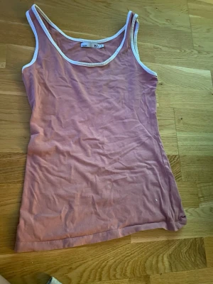 Rosa linnetopp från 157 - Säljer en enkel och snygg rosa linnetopp från 157 med vita kanter runt hals och ärmhål. Toppen är ärmlös och har en klassisk, rak passform. Perfekt att matcha med jeans eller shorts för en avslappnad look.