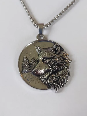 Halsband med vargmotiv i silverfärg - Snyggt halsband med ett runt hängsmycke i silverfärgad metall. Hänget har ett detaljerat motiv av ett varghuvud i 3D-effekt och bakgrund med granar. Kedjan är grov och matchar hängets färg. Materialet rostfritt stål.