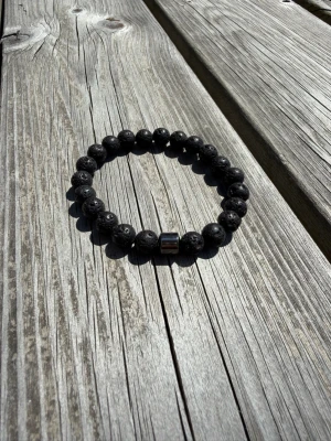 Svart armband med lavapärlor - Snyggt armband med svarta lavapärlor och en metallisk detalj. Pärlorna är runda och har en cool, porös yta som ger armbandet en unik look. Perfekt för dig som gillar stilrena accessoarer med lite edge.