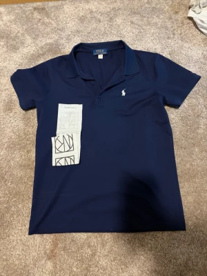 Marinblå pikétröja från Ralph Lauren - Klassisk marinblå pikétröja från Ralph Lauren med vit broderad logga på bröstet. Tröjan har korta ärmar, krage och knappar framtill. Perfekt för en stilren och sportig look. sitter som s/xs. Kvitto finns även till den