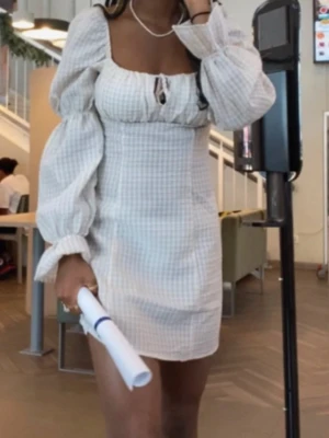 Vit kortklänning med puffärmar - Trendig vit kortklänning med rutigt mönster och puffiga långärmar. Klänningen har fyrkantig urringning, liten knytning framtill och markerad midja. Perfekt för dig som vill ha en söt och stilren look. Rutig och Off white färg 