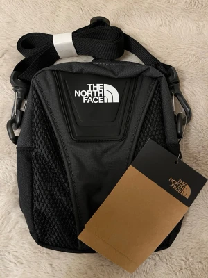 Svart axelväska från The North Face - Snygg svart axelväska från The North Face med justerbar axelrem och mesh-detaljer på sidorna. Väskan har en modern, sportig design med tydlig logga framtill och robust material som passar perfekt för att bära det viktigaste.