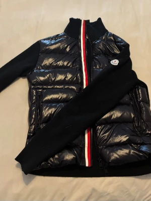 Svart dunväst från Moncler - Snygg svart dunväst från Moncler med glansig finish och quiltad design utan huva. Västen har en rödvit rand längs dragkedjan och två fickor med dragkedja. Klassisk Moncler-logga på bröstet och hög krage för extra stilpoäng. 
