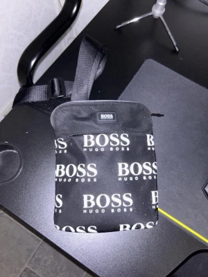 Hugo Boss Väska - Snygg svart axelväska från Hugo Boss med justerbar rem och tydligt logomönster i vitt över hela väskan. Väskan har en fyrkantig form och dragkedja upptill. Perfekt för att bära det viktigaste och ger en trendig streetwear-look. Säljs för brist på användning.  Fraktas endast och köparen står för frakt.